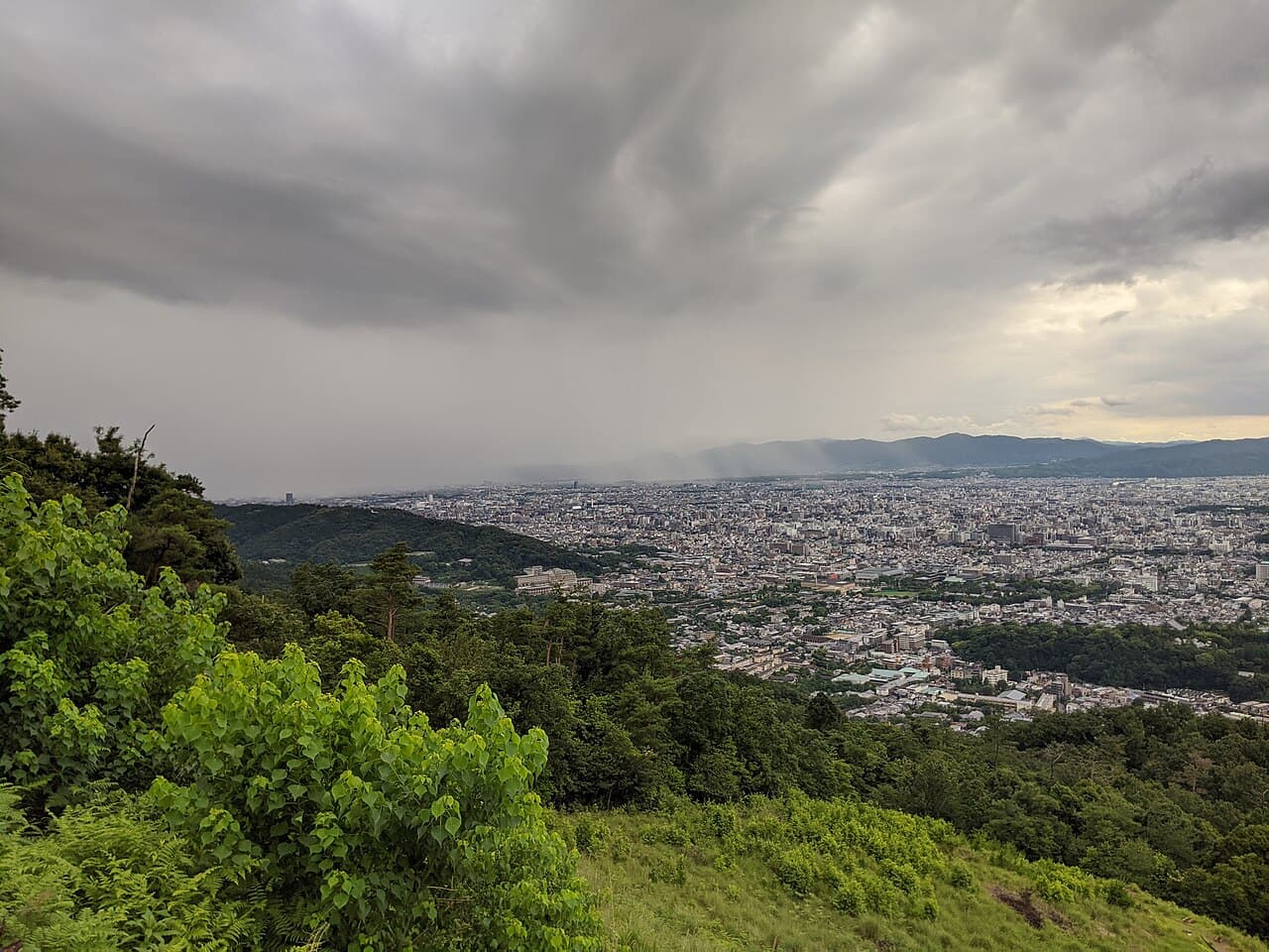 Kyoto seen from Daimonji hill under heavy grey clouds: green trees and grass in the foreground, then a dense spread of low- and mid-rise buildings running to mountains on the horizon